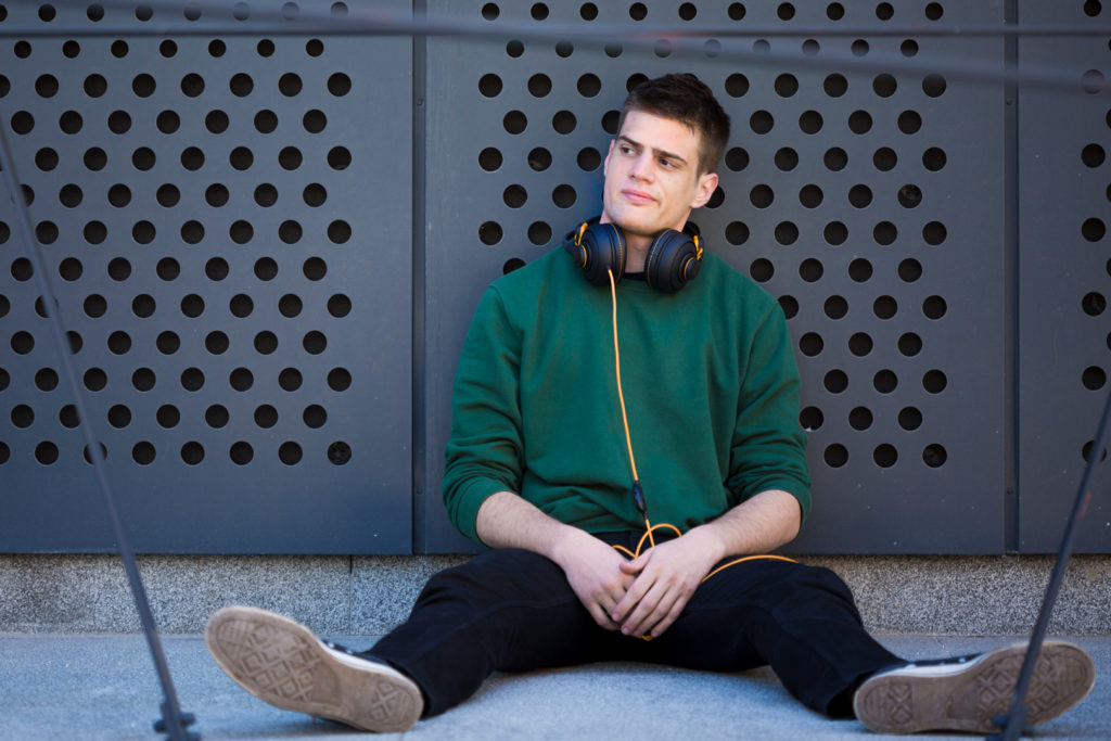 Männlicher Jugendlicher mit Kopfhörern um den Hals sitzt auf dem Boden, angelehnt an eine Wand, und schaut desillusioniert zur Seite